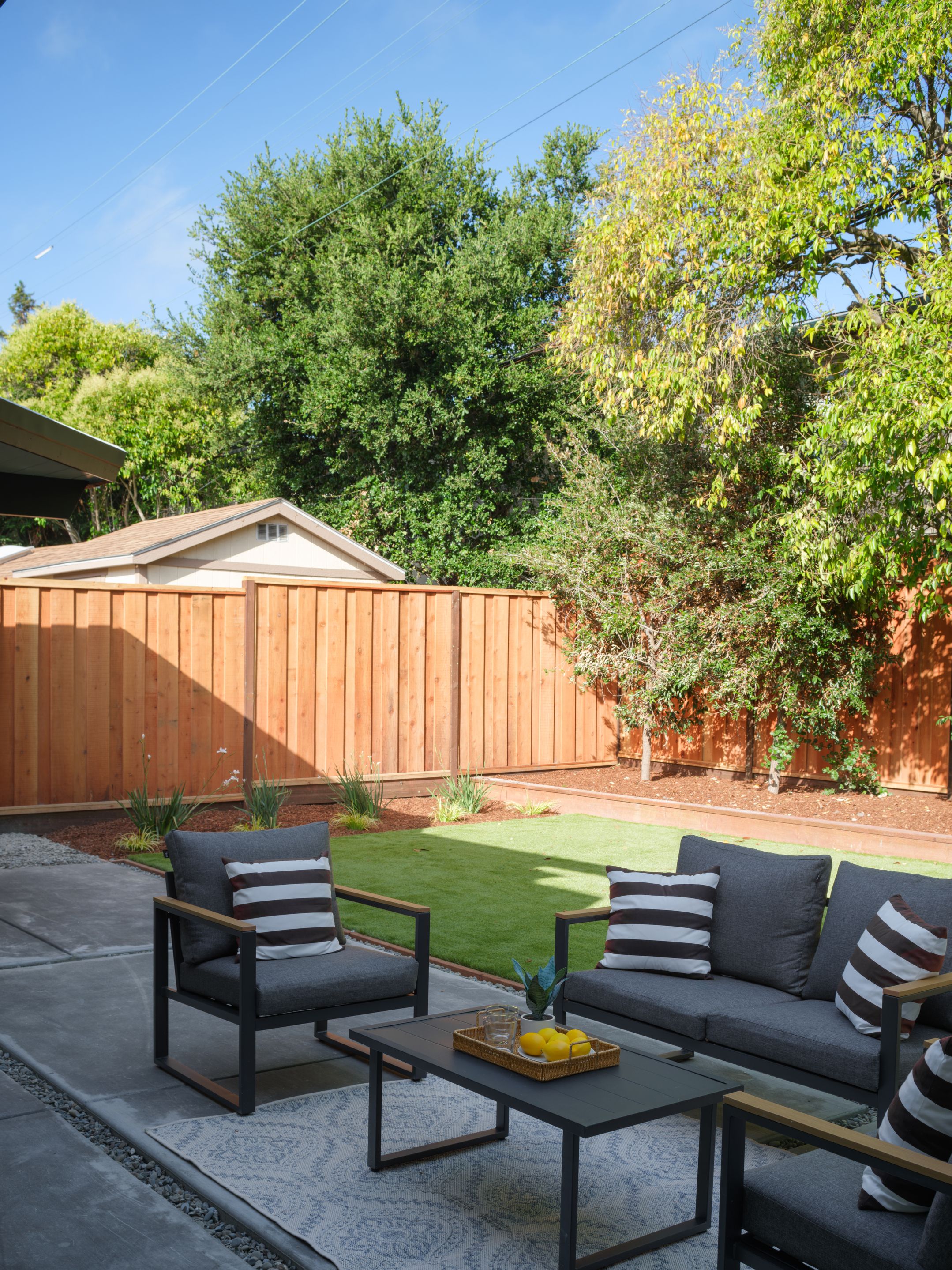 Outdoor patio lounge with gray sofa, striped pillows, cedar fence, and bright lawn for indoor-outdoor staging