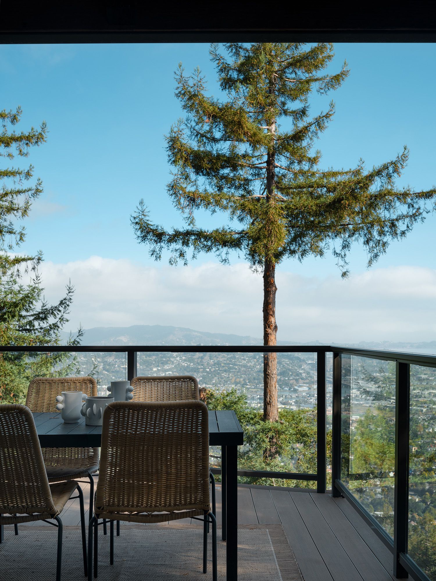 Covered deck dining area with woven chairs, black table, glass railing, and hillside views for summer entertaining