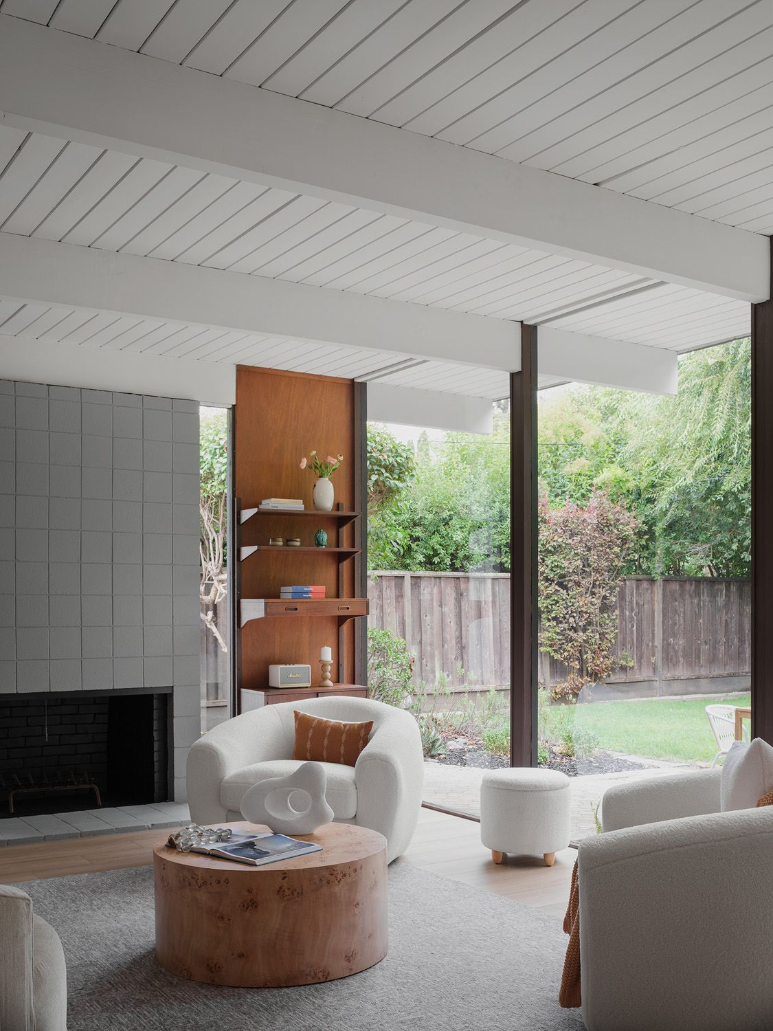 Sunlit mid-century living room with cream seating, burl wood table, and garden-facing glass walls showing coastal style led by light