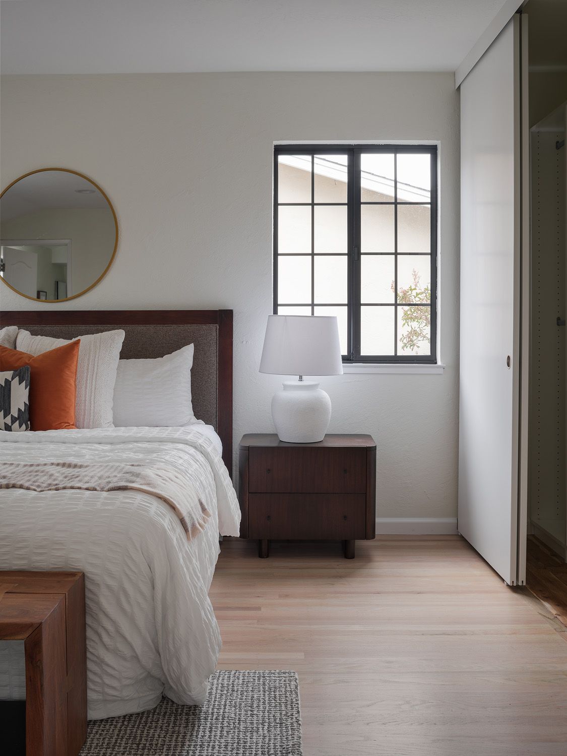 Smoothed white bedding, walnut nightstand, ceramic lamp, brass mirror, and bright window in a staged bedroom