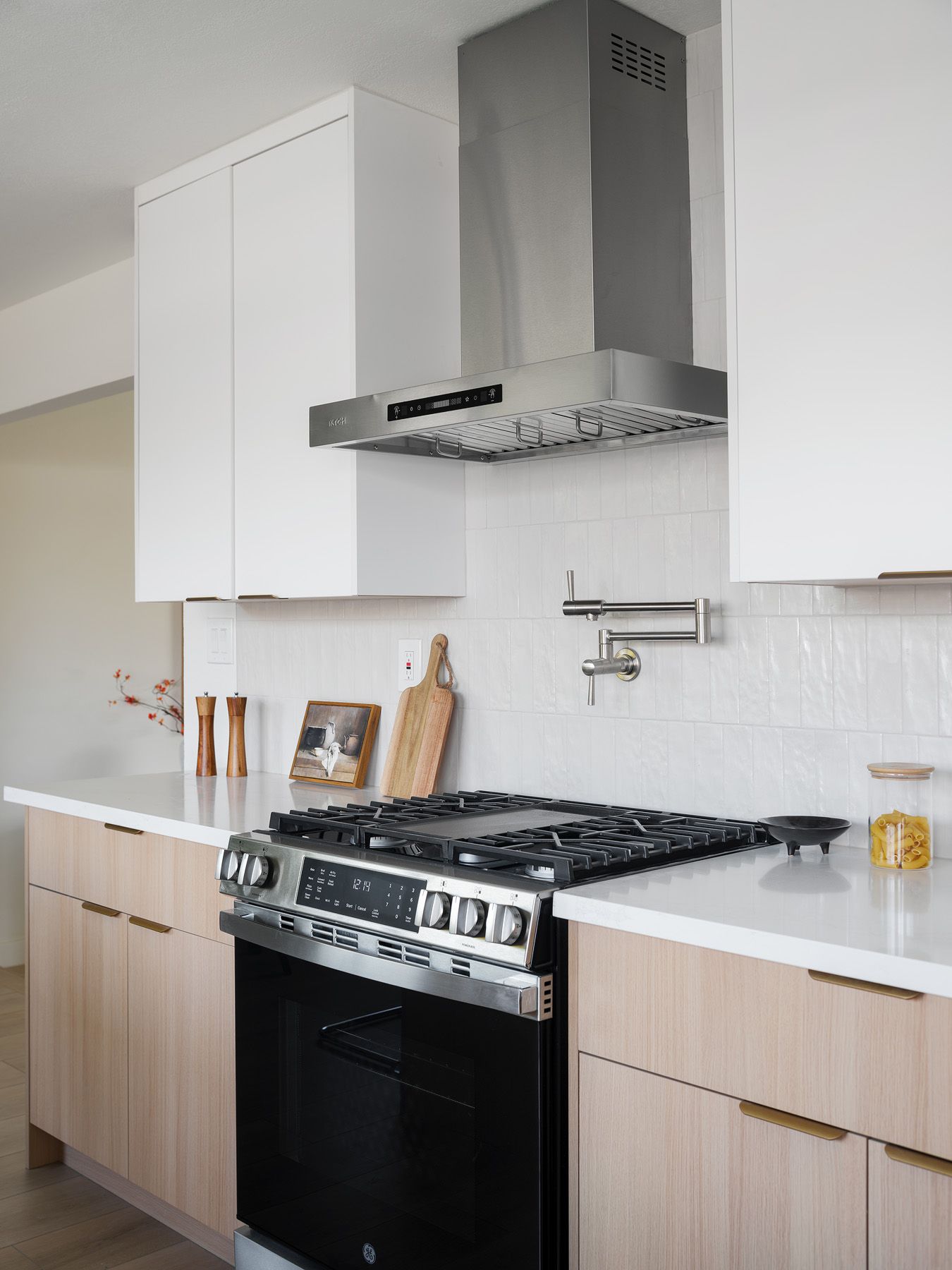 Light oak cabinets with brass pulls, white tile and a stainless range hood illustrate hardware coordinated with nearby metals
